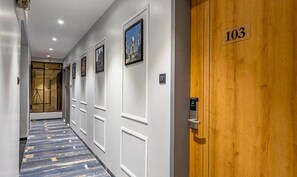 Un pasillo con una puerta de madera con el número 103, paredes blancas y cuadros enmarcados.