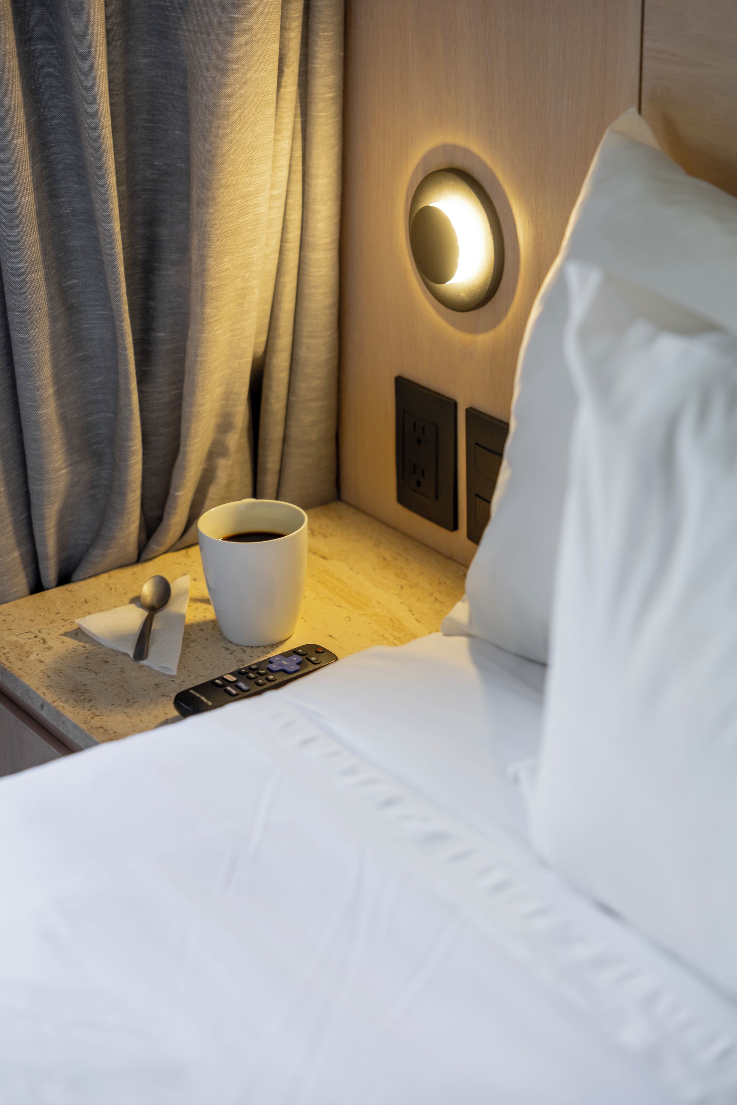 A hotel room with a neatly made bed, a bedside table with a remote control, a cup of coffee, and a wall-mounted light fixture.