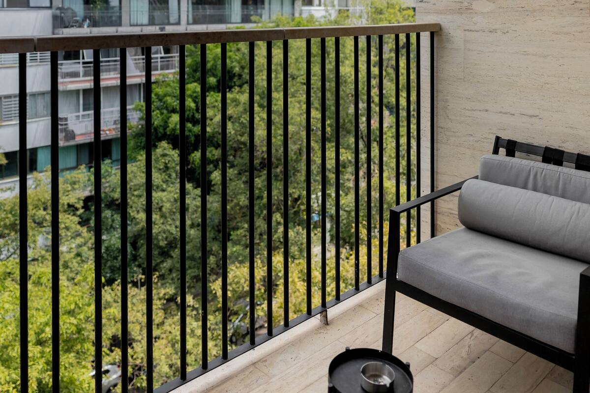 A balcony with a grey sofa, a small table with a bowl, and a railing. A modern building with balconies is visible in the background.