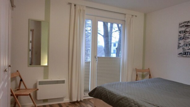 A bedroom with a bed, a radiator, a window with curtains, a chair, and a wall-mounted mirror.