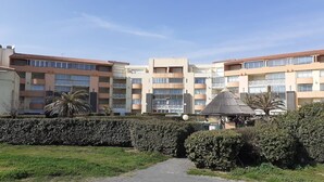 Un immeuble résidentiel à plusieurs étages, doté de balcons, d’une cour intérieure avec une structure recouverte de chaume, et de haies bien entretenues.