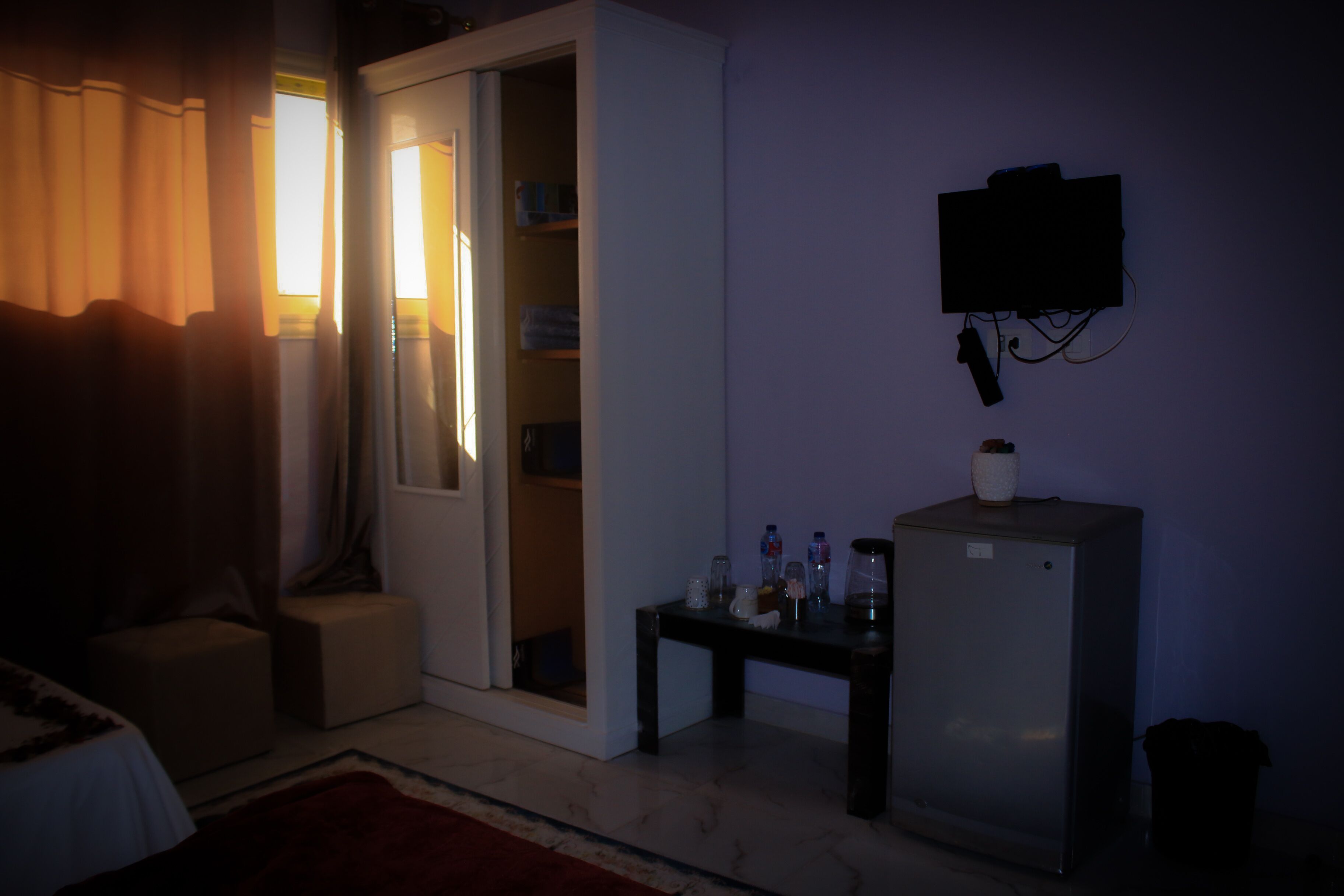 Una habitación poco iluminada con una puerta cerrada, una pequeña mesa con botellas y un refrigerador.