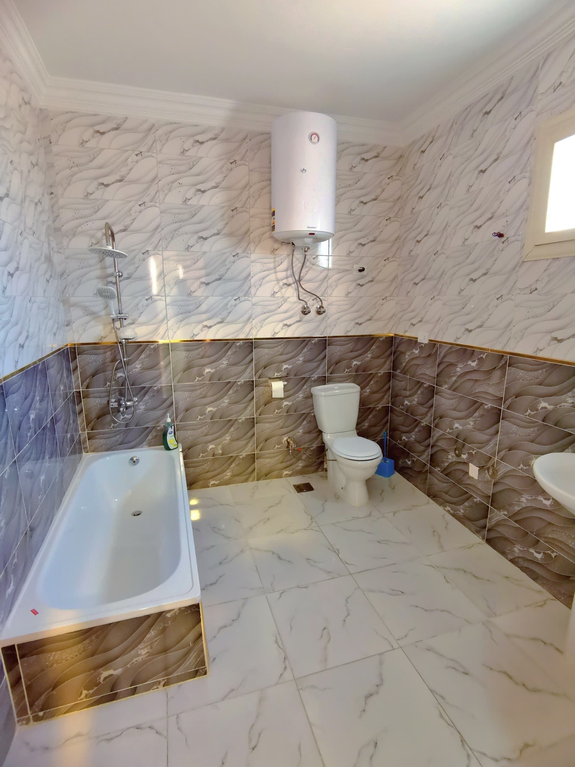 Un baño con bañera, inodoro y lavabo, con azulejos de aspecto marmolizado en paredes y suelo.