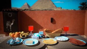 Mesa dispuesta con alimentos para el desayuno, como frutas, pan y bebidas, con una pirámide y cielo despejado al fondo.