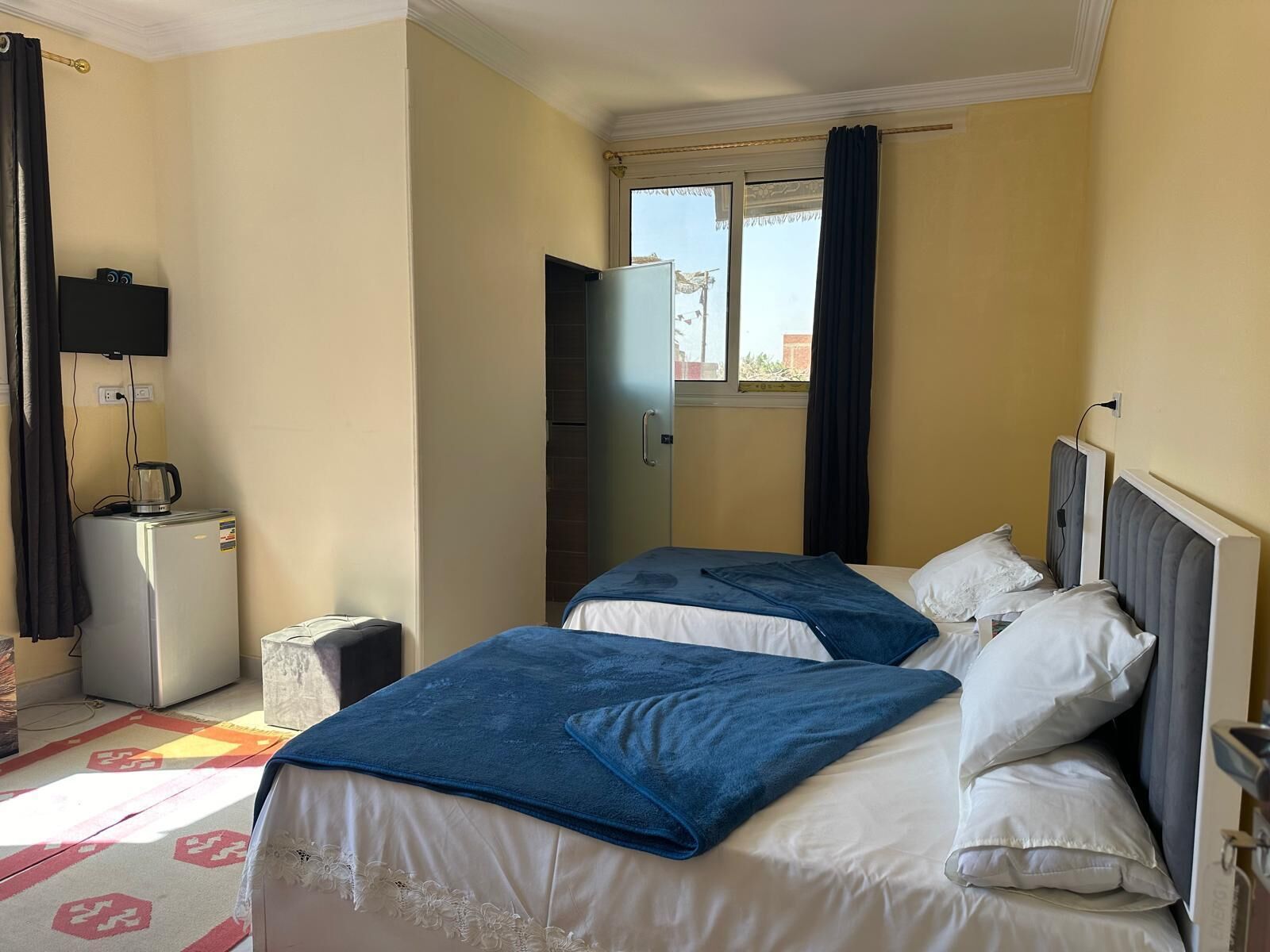 A hotel room with two beds, a small kitchenette, a TV, and a window with curtains.