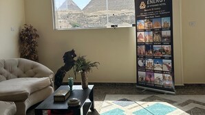 Un vestíbulo de hotel con un amplio ventanal que ofrece vistas a las pirámides, un sofá, una mesa de centro con objetos decorativos y un expositor promocional del hotel.