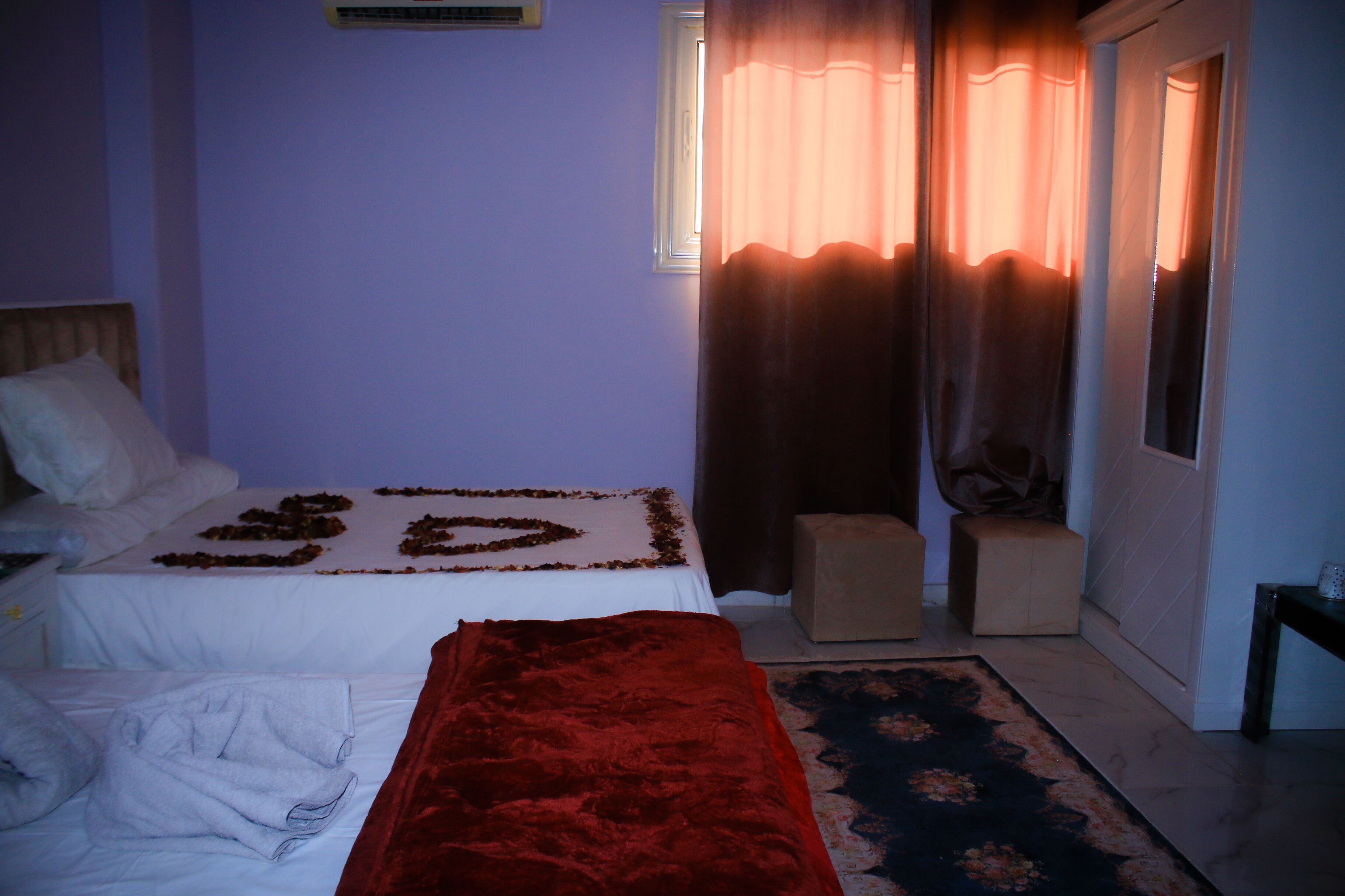 Quarto com cama, manta vermelha, um banquinho pequeno e tapete estampado.