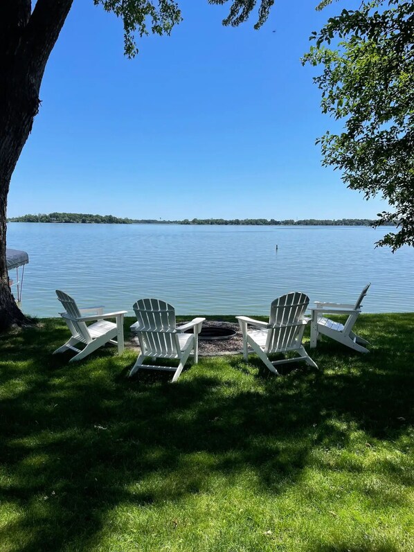 A lakeside view with two Adirondack chairs facing a calm lake, surrounded by lush green grass and a large tree.