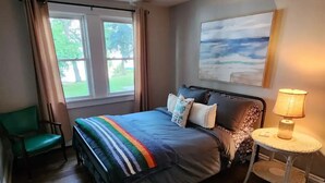 Une chambre à coucher avec un lit, un ventilateur de plafond, une fenêtre avec des rideaux, une chaise, une table avec une lampe et un tableau au mur.