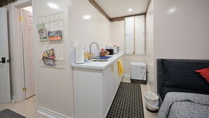 Een kleine keuken met een magnetron, spoelbak en een rek voor het afdrogen van vaat. Een kleine koelkast is zichtbaar, en er ligt een vloerkleed met een patroon op de vloer.