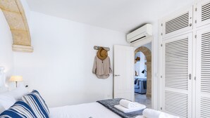 Et hotelværelse med en seng, et natbord, en lampe, en hat på væggen og udsigt til et andet værelse gennem en åben dør.