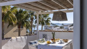 En terrasse med et hvidt spisebord og hvide stole, omgivet af palmer og udsigt til havet samt bygninger i baggrunden.