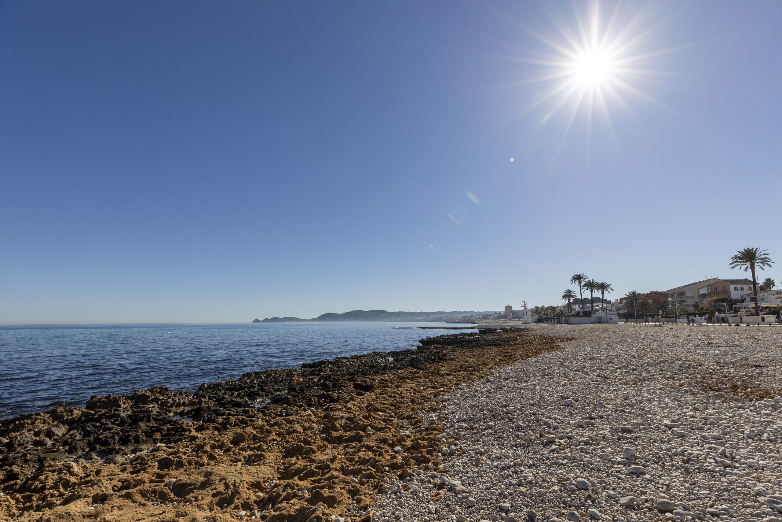 A beach with pebbles and sand, a clear sky, and buildings in the background.