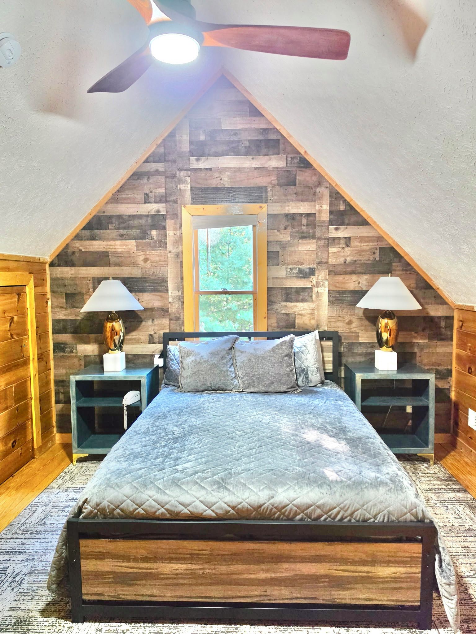 A bedroom with a sloped ceiling, wooden wall paneling, a bed with a quilted bedspread, two bedside tables with lamps, and a window.