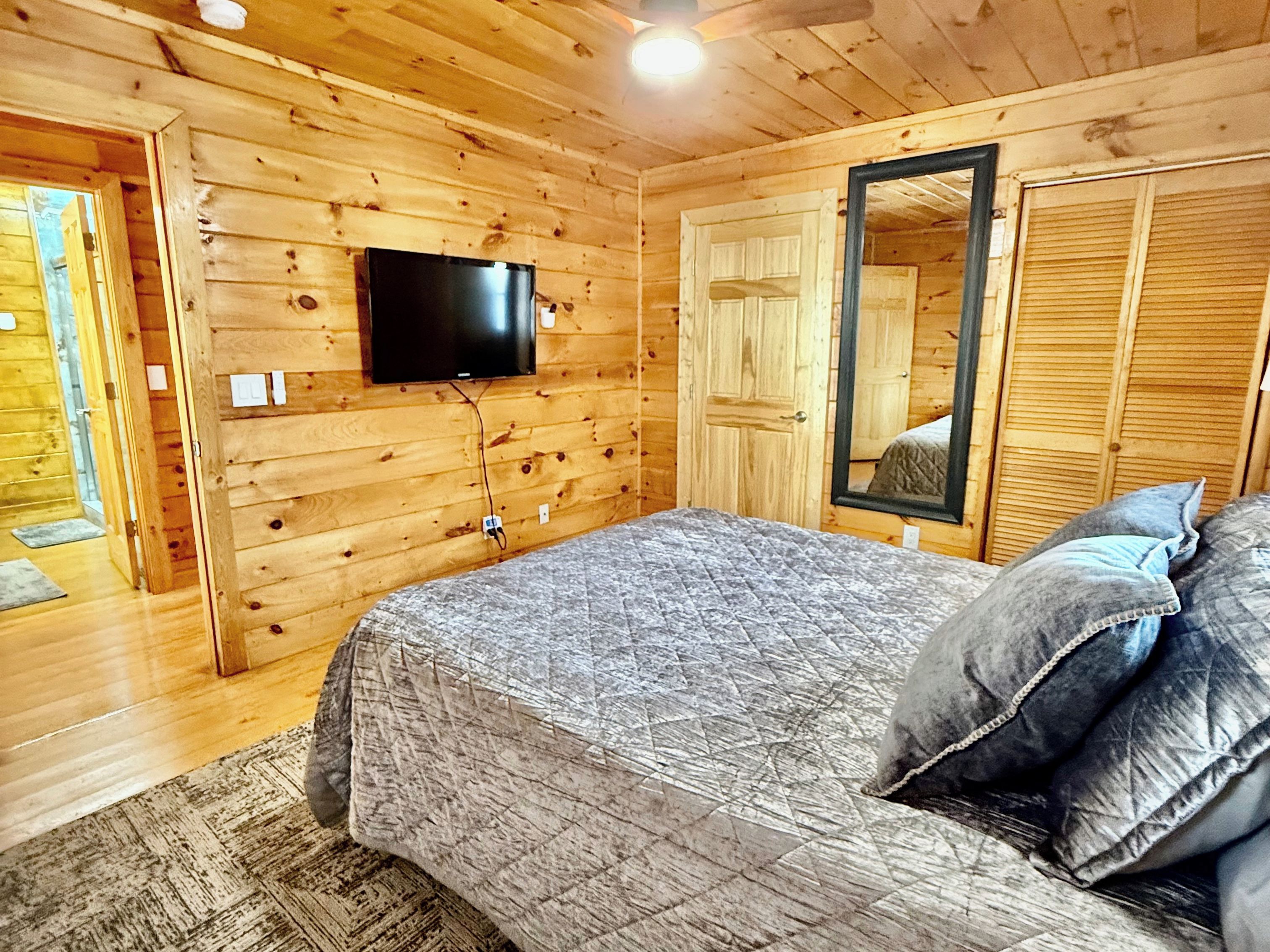 A wooden-paneled bedroom with a bed, a flat-screen TV mounted on the wall, and a large mirror.