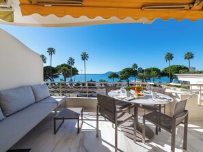 Terraço com um sofá branco, mesa de jantar posta com pratos, copos e uma garrafa, com vista para um mar azul límpido, palmeiras e uma montanha ao longe.