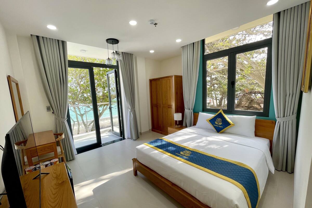 A hotel room with a bed, a desk, and a wardrobe. There is a window with curtains and a view of the outside.