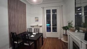 En matplats med ett bord och stolar, ett element, ett fönster med gardiner och en växt på en piedestal.
