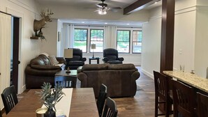 A spacious living room with a dining area, featuring a ceiling fan, large windows, and a mounted deer head.