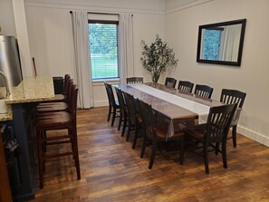 A dining room with a long wooden table, dark chairs, a potted plant, and a window with blinds.