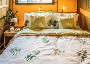 Ein Bett mit blattgemusterten Bettbezügen, eine Nachttischlampe und ein Topfpflanze auf einem Holznachttisch.