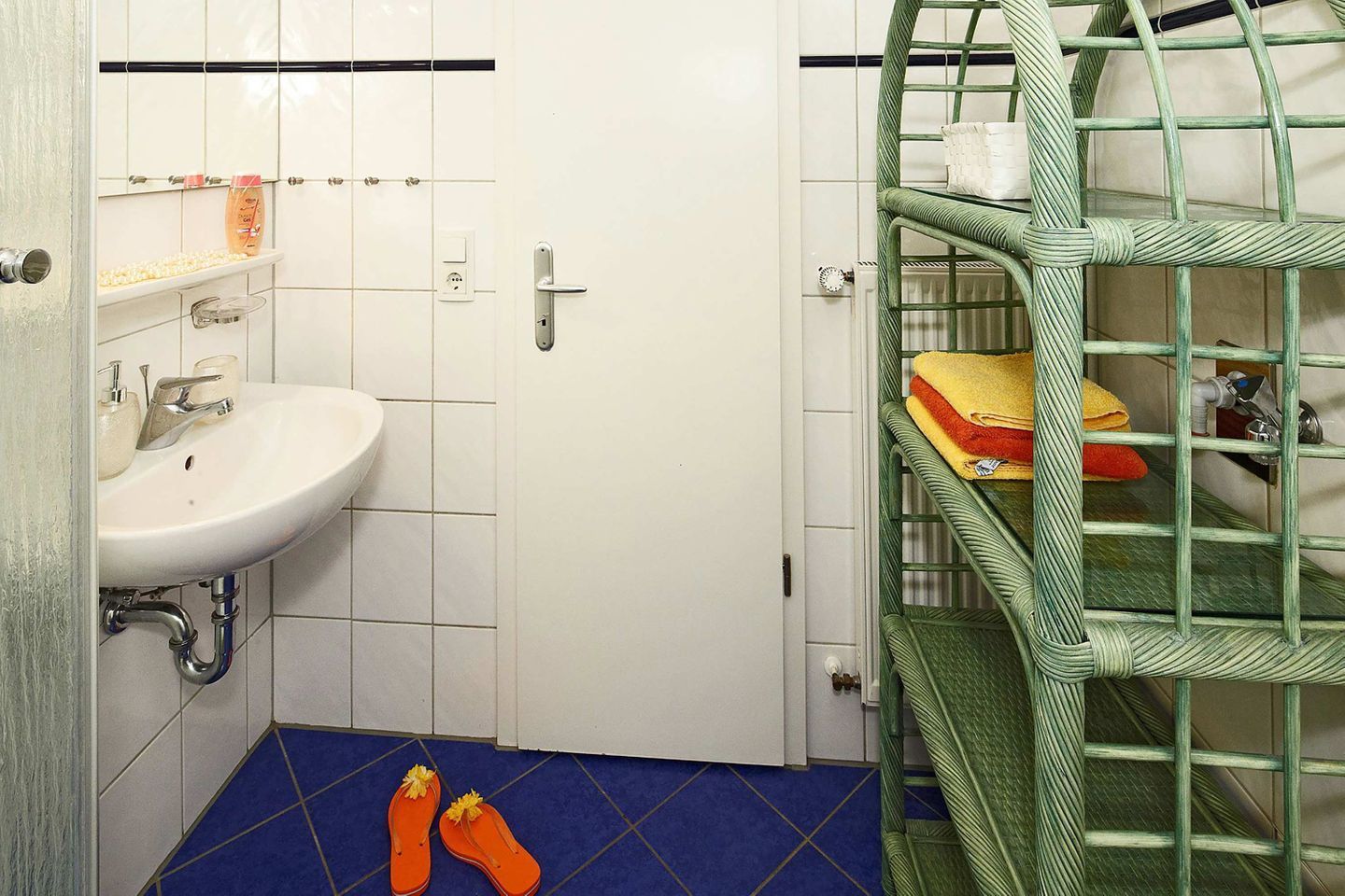 Un baño con un lavamanos blanco, piso de azulejos azules y una estantería metálica verde.