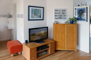 A living room with a TV, wooden cabinet, red ottoman, and wall art. - Traumhafte Ferienwohnung 50 m von der Seebrücke in Koserow, mit Wlan und Balkon - Seeblick Anlage (Koserow)