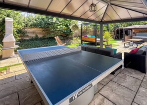 An outdoor ping pong table with a pergola overhead, surrounded by outdoor furniture and a barbecue area.