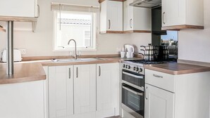 A compact kitchen with white cabinets, a stainless steel sink, a stove, and a window with blinds. - 3 bedroom accommodation in Chapel St Leonards, Skegness (Chapel St Leonards, Skegness)