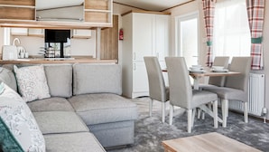 A modern living room with a sofa, dining table, and chairs, a kitchen area with cabinets and a stove, and a large window with curtains. - 3 bedroom accommodation in Chapel St Leonards, Skegness (Chapel St Leonards, Skegness)