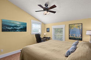 A bedroom with a bed, a desk, a chair, a ceiling fan, and artwork on the walls.