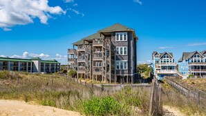 A beachfront hotel with multiple balconies, a sandy beach in the foreground, and a clear blue sky. - Sand Castle 4: Oceanfront Condo, Community Pool, Ocean Views from Deck (Kill Devil Hills)