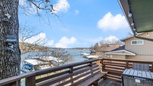 Eine Holzterrasse mit Geländer, Blick auf einen See, wo Boote angedockt sind, und ein Haus im Hintergrund.