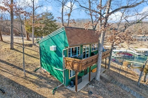 A small, green, two-story cabin with a wooden deck and a balcony, surrounded by trees and a dock. - Fishing, Boat Slip, Morning Coffee, Picturesque View (Sunrise Beach)