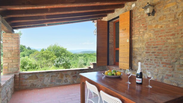 Un tavolo di legno con una ciotola di frutta e bicchieri da vino su un balcone con un muro in pietra e travi di legno.