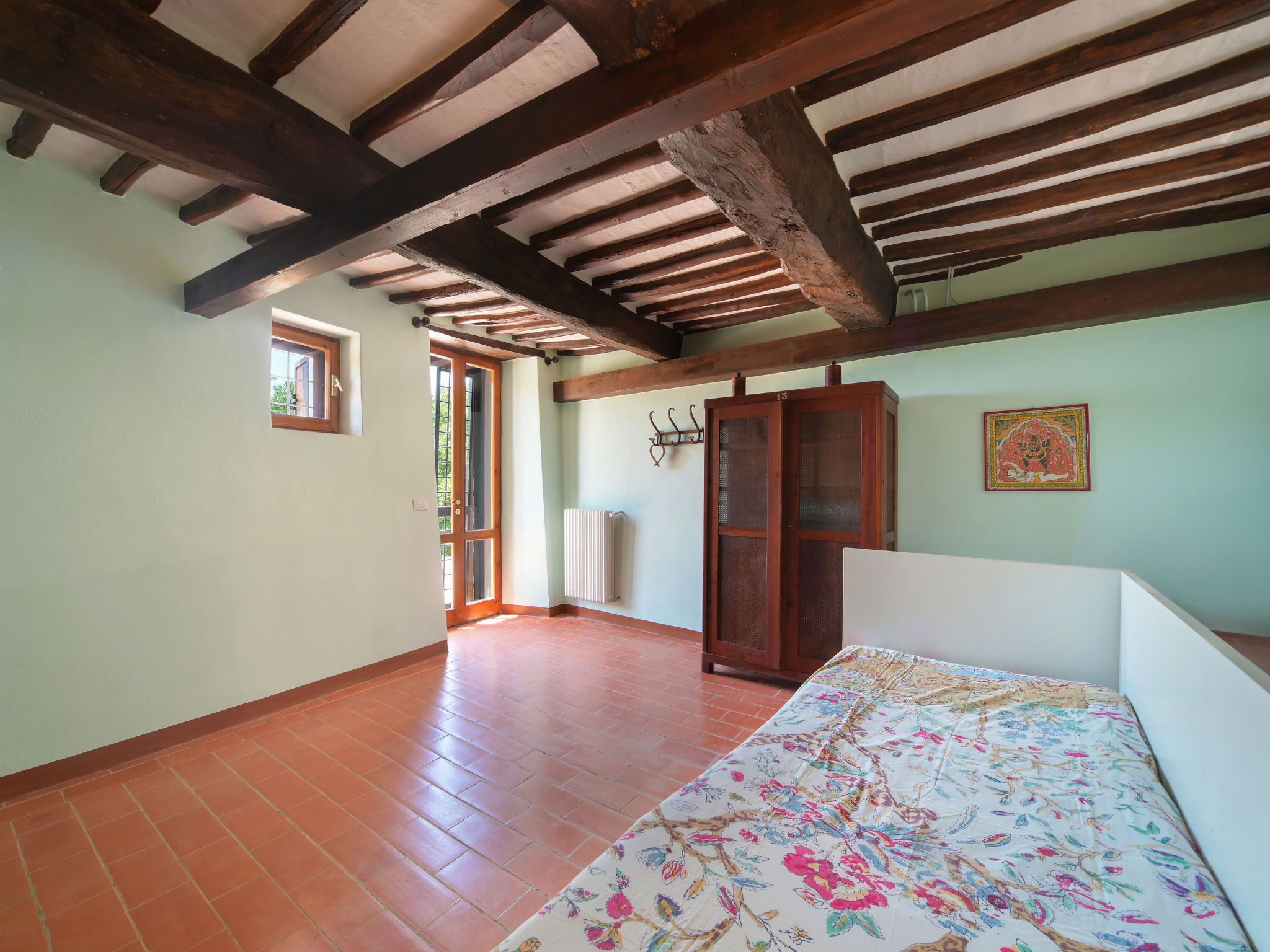 A room with exposed wooden beams, a tiled floor, a bed with a floral bedspread, a wardrobe, and a radiator.
