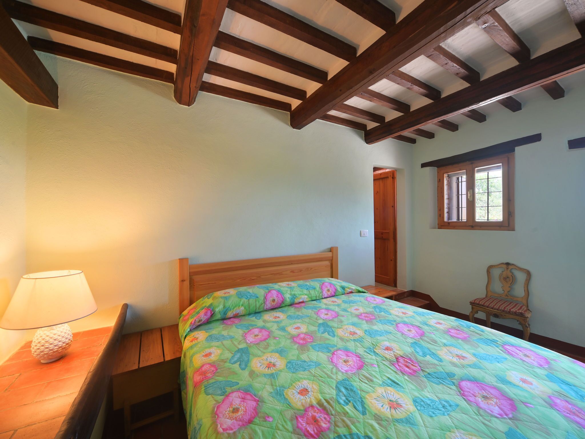 Quarto com cama de madeira, mesa de cabeceira, luminária e janela com grades.