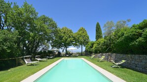 Una piscina rettangolare con una parete in pietra da un lato e un'area verde con lettini dall'altro.