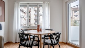 A dining area with a round table, four chairs, a vase, and a view of buildings through large windows. - Tasteful Iselin 2BR w/ Storage, steps to Central Market, by Blueground (Basel)