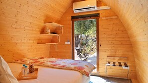 A cozy wooden room with a bed, a small table with a bottle and a tray, a bench, and an air conditioning unit.