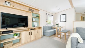 A modern living room with a flat-screen TV, a sofa, a dining table, and a chair. There are wooden shelves with decorative items and a window with curtains.
