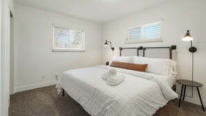 A bedroom with a bed, two bedside lamps, a small table, and two windows with blinds. - DenverStays: 5BR Stylish and Modern Home w/Garage, great for pets and families! (Arvada)