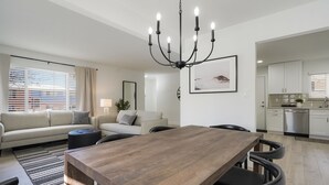 A modern living room with a wooden dining table, two sofas, a chandelier, and a kitchen in the background. - DenverStays: 5BR Stylish and Modern Home w/Garage, great for pets and families! (Arvada)