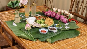 A dining table with a banana leaf placemat, a vase with flowers, and a variety of food items including rice, vegetables, and a drink. - Cybele Sunset Hotel Phu Quoc (Phu Quoc)