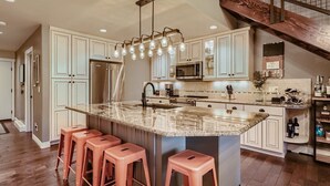 A kitchen with white cabinets, a granite island with stools, stainless steel appliances, and a wooden staircase.
