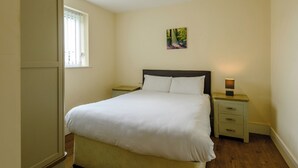 A bedroom with a bed, a nightstand, a bedside table, a wardrobe, and a window. - Ground Floor Apartment (Pet) (Ingoldmells, Skegness)