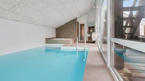 An indoor swimming pool with clear water, a stone wall, and a view of the outside through large windows.