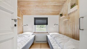A small, well-lit room with two single beds, wooden ceiling, and a window with blinds.