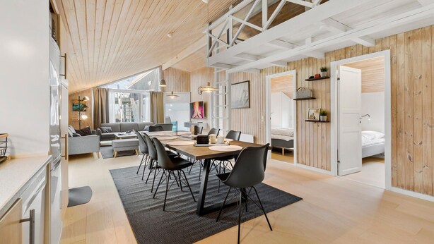 A spacious living area with a high ceiling, wooden beams, a dining table with chairs, a sofa, and a bed in the adjacent room.