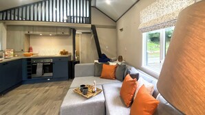 A modern interior with a kitchen, a sofa with orange cushions, and a view of greenery outside. - Relax and unwind in the perfect hideout. (Snowdonia)